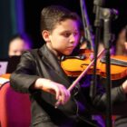 Imagem mostra menino de cerca de dez anos usando terno preto e tocando um violino