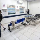 Em uma sala ampla de uma unidade de saúde, um homem está varrendo o chão, enquanto outro manuseia equipamentos. Há macas e cadeiras no espaço.