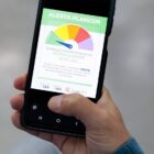 Imagem mostra mão de um homem segurando um celular. Na tela, há uma imagem da identidade visual do Plancon, que simula um velocímetro, com o ponteiro no estágio verde