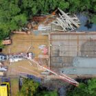 Imagem aérea mostra a laje de uma ponte sendo concretada. Perto da área onde será a ponte, há um caminhão betoneira com uma grande mangueira que leva o cimento até o local, onde há armações em ferro. Há pessoas trabalhando.
