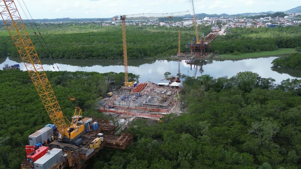 Imagem aérea mostra guindastes em outros equipamentos em obra próxima a rio e à área de vegetação
