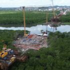 Imagem aérea mostra guindastes em outros equipamentos em obra próxima a rio e à área de vegetação