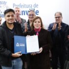 Um homem e uma mulher seguram juntos um documento e o mostram para a câmera. Ao lado deles, há outros homens e mulheres que sorriem e aplaudem