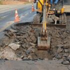 Imagem mostra máquina escavadeira escavando buraco em rua de asfalto sinalizada com cones
