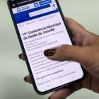 Mão feminina com unhas pintadas de marrom segura um celular com tela mostrando página da Conferência Municipal de Saúde no site da Prefeitura de Joinville