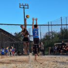 Imagem mostra dois jovens em uma quadra de vôlei de areia. Os dois estão próximos à rede, com braços ao alto para alcançar a bola. Ao redor da quadra, outras pessoas assistem ao jogo