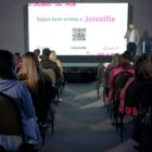 Um homem está no palco, em pé e segurando um microfone, à frente de um painel que diz sejam bem-vindos a Joinville. Na sala, há várias pessoas sentadas assistindo à apresentação