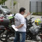 Homem de camiseta branca conversa com motociclista. Ao fundo, há motocicletas