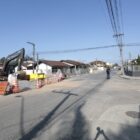 Imagem mostra uma rua asfaltada com homem passando em bicicleta. Em uma lateral da via, há uma máquina retroescavadeira, além de brita, cones e placas.