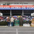 Imagem mostra duas mulheres e um homem fardado com roupas do exército em pé, em frente a uma grande pilha de roupas e cobertores