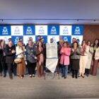 Homens e mulheres estão em pé em um palco. Há uma placa metalizada que marca a inauguração da Casa Abrigo Viva Rosa no centro, e as pessoas sorriem e aplaudem
