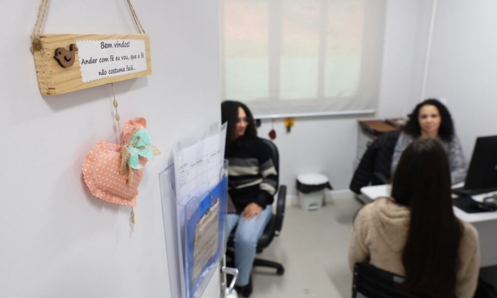 Imagem mostra porta branca entreaberta. Dentro do local, há três mulheres sentadas, uma de costas para a câmera