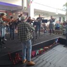 Imagem mostra pessoas, algumas vestidas de xadrez, tocando instrumentos musicais em um ambiente decorado de festa julina