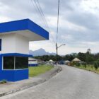 Imagem mostra uma guarita pintada de azul e branco ao lado de uma estrada que leva a uma construção. Há árvores e gramado no entorno.