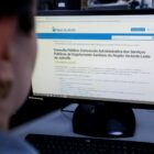 Mulher com o rosto parcialmente visível e desfocado observa tela de computador em página do site da Águas de Joinville sobre consulta pública