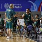 Jogador de basquete em cadeira de rodas está sentado na cadeira e cumprimenta um homem em pé em uma quadra de basquete. Ao redor deles, há outros jogadores.