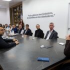 Em uma sala ampla, há uma grande mesa onde há uma mulher loira sentada na ponta, enquanto homens e mulheres ocupam os outros assentos, enquanto olham para ela