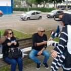 Pessoa vestida de mosquito da dengue conversa com duas pessoas sentadas no banco de uma praça. Uma mulher de colete da vigilância acompanha a conversa.