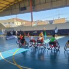 Em uma quadra de esportes, há crianças sentadas em cadeiras de rodas aprendendo a como jogar basquete