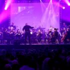 Imagem mostra músicos vestidos de preto em um palco com iluminação roxa