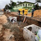 Imagem mostra uma grande vala em uma rua. Dentro dela, há grandes caixas de concreto e homens trabalhando