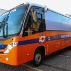 Micro-ônibus laranja com detalhes em azul está estacionado em um pátio aberto