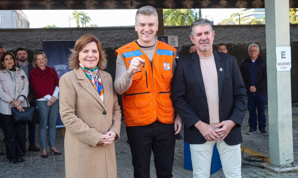 Homem loiro de colete laranja segura uma chave e posa para a foto sorrindo. Ao lado dele, estão uma mulher e um homem também sorrindo
