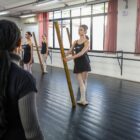 Bailarinas com meias finas, sapatilhas e figurino preto ensaiam em uma sala. Perto delas, uma mulher observa