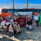 Alunos de uma fanfarra estão vestidos com roupas vermelhas e detalhes em dourado. Eles estão em pé, segurando os instrumentos, em frente a um ônibus em um pátio
