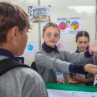 Imagem mostra uma adolescente apontando para um cartaz em uma parede. Atrás dela, há outra estudante e à frente um menino prestando atenção à explicação