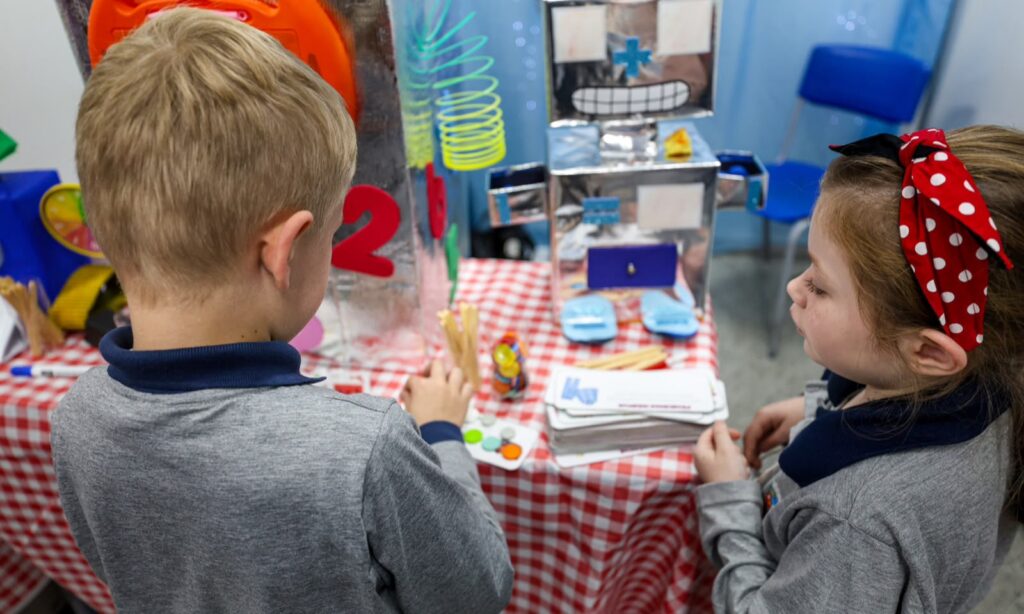 A imagem mostra duas crianças de cerca de 4 anos, um menino e uma menina, manuseando objetos em uma mesa com toalha xadrez