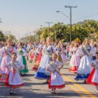 Imagem mostra meninas e mulheres com figurino típico alemão desfilando em uma rua pavimentada. Às margens da rua, pessoas acompanham o desfile