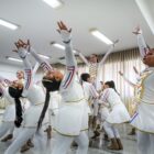 Adolescentes vestidas com figurino branco ensaiam a coreografia em uma sala ampla