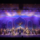Dezenas de bailarinos usando figurino de balé clássico estão parados em pose de coreografia em um grande palco iluminado com tons de rosa e roxo