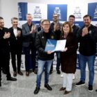 Homens e mulheres estão em pé, em frente a um painel da prefeitura, aplaudindo e sorrindo. No centro, um homem e uma mulher seguram juntos um documento