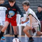 Dois adolescentes estão em movimento em uma quadra de futsal. Jogadores de times diferentes, eles correm em direção à bola
