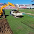 Imagem mostra uma máquina retroescavadeira despejando terra e grama em um caminhão caçamba. Os veículos estão no gramado de um estádio de futebol.