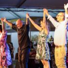 Duas mulheres e dois homens estão de mãos dadas e braços levantados em um palco. Eles sorriem para a plateia