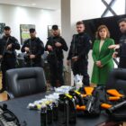 Em uma sala ampla, há quatro homens uniformizados e armados com uma arma longa junto ao corpo. Ao lado deles, há uma mulher e outro homem que fala enquanto aponta para uma mesa com outras armas e sprays de pimenta