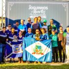 Imagem mostra atletas e técnicos em frente a um painel dos Joguinhos Abertos de Santa Catarina. No centro, três mulheres seguram uma bandeira de Joinville e um troféu