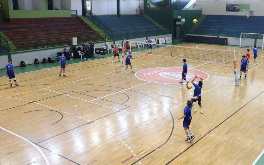 Imagem mostra quadra de vôlei com atletas vestidos de azul treinando com bola