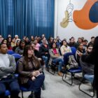 Imagem mostra uma sala repleta de pessoas sentadas em cadeiras. À frente, um homem vestido de preto fala e gesticula para as pessoas.