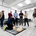 Em uma sala de aula, um homem está ajoelhado fazendo massagem cardíaca em um boneco. Ao redor, homens e mulheres assistem à cena