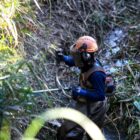 Homem equipado com luvas, capacete e viseira utiliza uma roçadeira nas margens de um rio