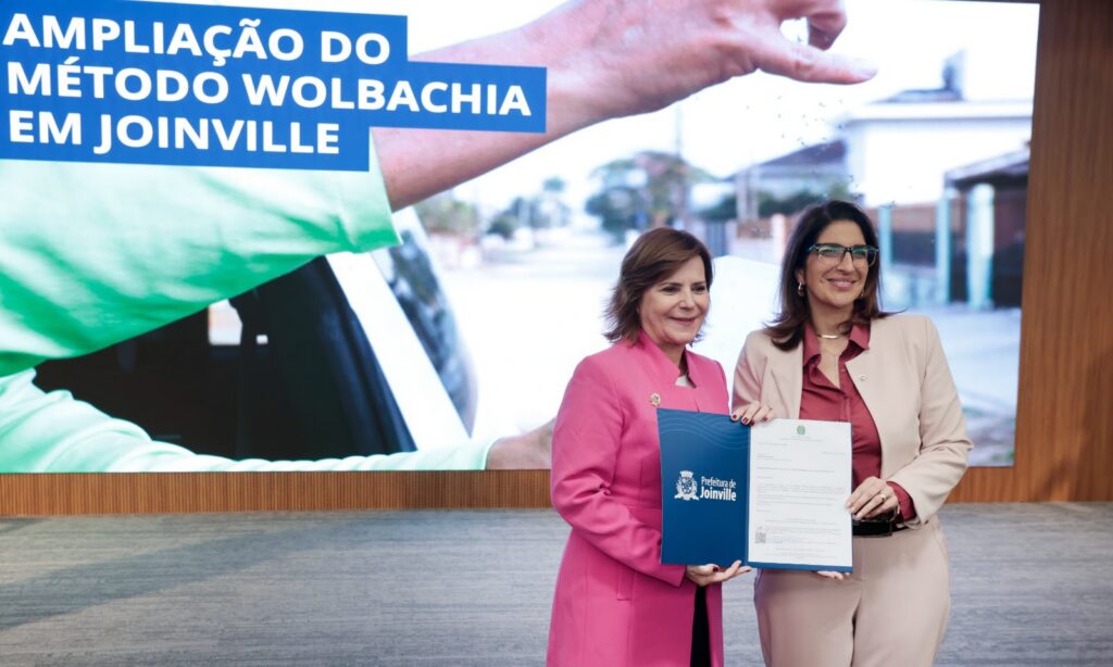 Duas mulheres seguram juntas um mesmo documento, mostrando para a plateia. Elas estão juntas, em frente a um palco