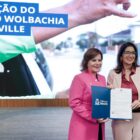 Duas mulheres seguram juntas um mesmo documento, mostrando para a plateia. Elas estão juntas, em frente a um palco