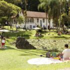 Imagem mostra uma casa de dois andares branca ao fundo, onde fica o Museu de Arte de Joinville. Em primeiro plano, está um lago e um grande gramado onde há crianças e adultos