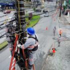 Imagem área mostra dois trabalhadores com roupas de proteção e capacete em cima de duas escadas, cada uma de um lado de um poste. No chão, há outros trabalhadores