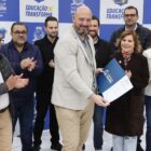 Em frente a um grande painel da Prefeitura, homens e mulheres estão em pé, sorrindo e aplaudindo. No centro, um homem e uma mulher seguram juntos um documento