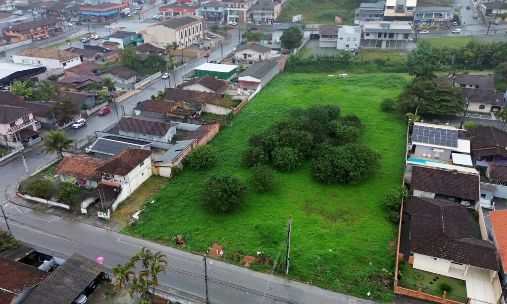 Imagem aérea mostra um grande terreno com gramado verde e algumas árvores. Nas laterais do terreno, há várias casas e, à frente, uma rua asfaltada.